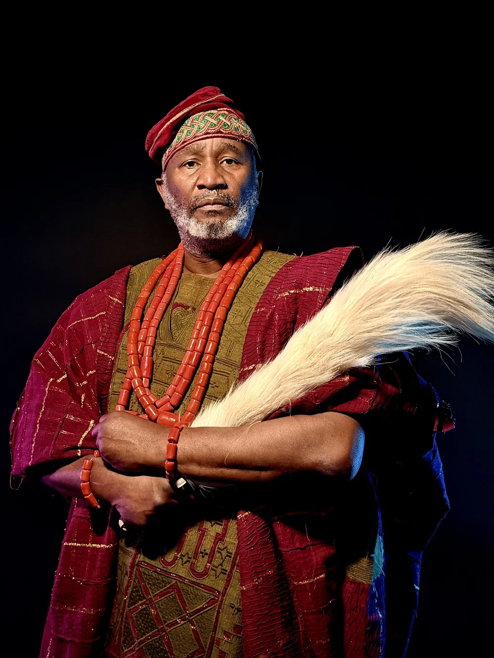 An elder in traditional chief's regalia with coral beads, photographed against black