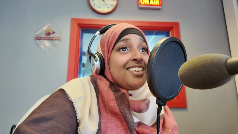 A young presenter in a hijab and headphones smiling at the microphone under a glowing on-air sign